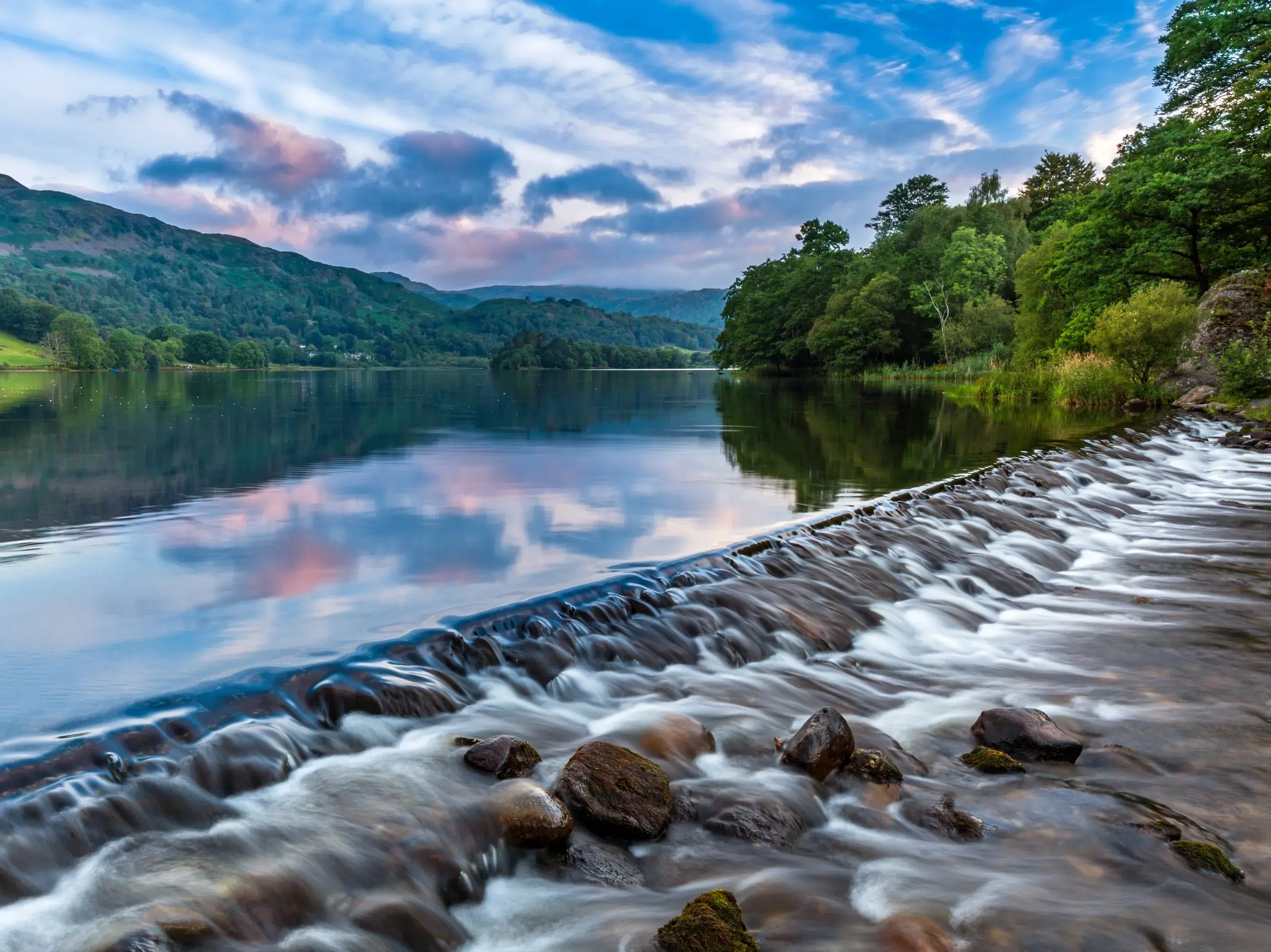 The River Rothay flowing over the weir at Grasmere Lake towards Rydal. A lovely summer morning sky over the Barrowdale fells in the distance scaled